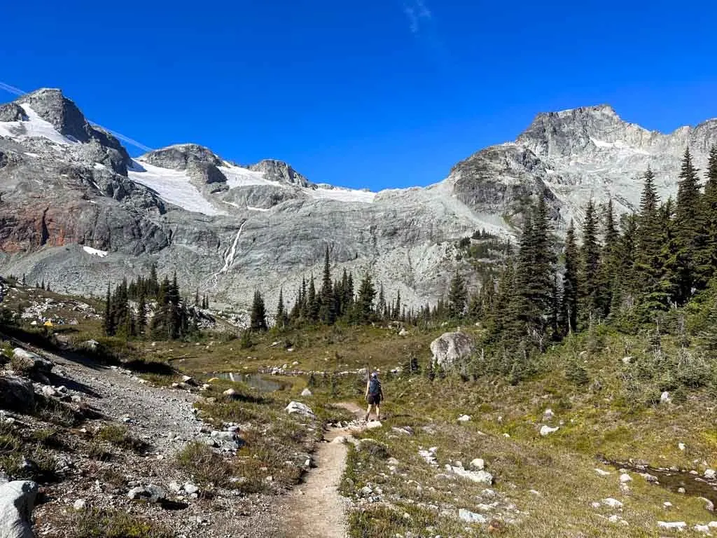 A hiker on an alpine trail with a waterfall and a glacier in the background