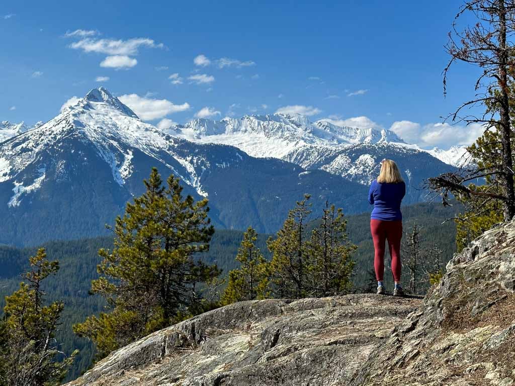 A woman stands on a bluff near Brohm Lake with snowy mountains in the distance. Get everything you need to know to hike at Brohm Lake