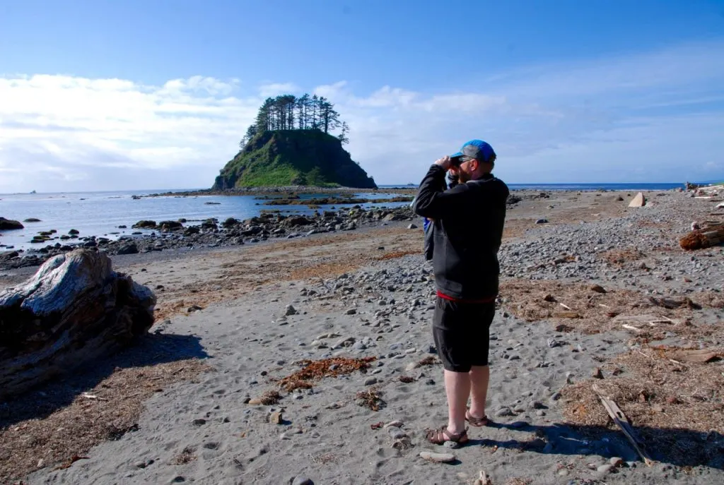 A hiker stands near Cape Alava and looks through binoculars with Tskawahyah Island in the background