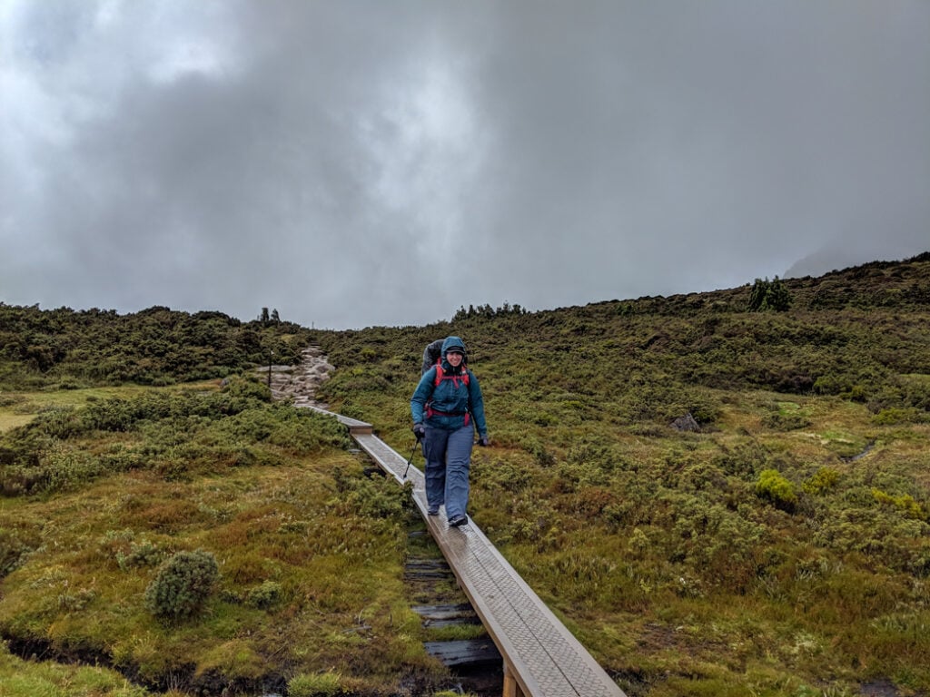 A hiker walks along a boardwalk on the Overland Track in Tasmania. It is raining and she wears a rain jacket and rain pants.