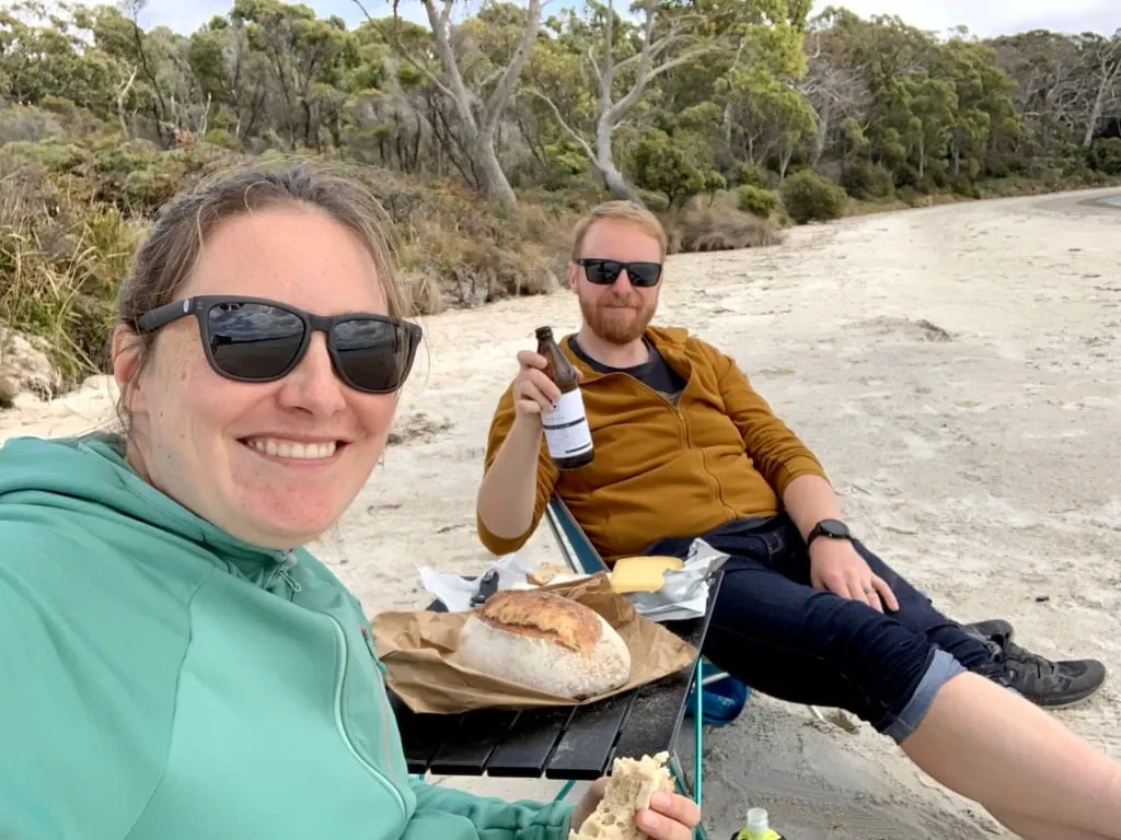 Two people sit in camp chairs and eat off a small folding table on a beach. The table is from Amazon, which is a place to save money on hiking gear