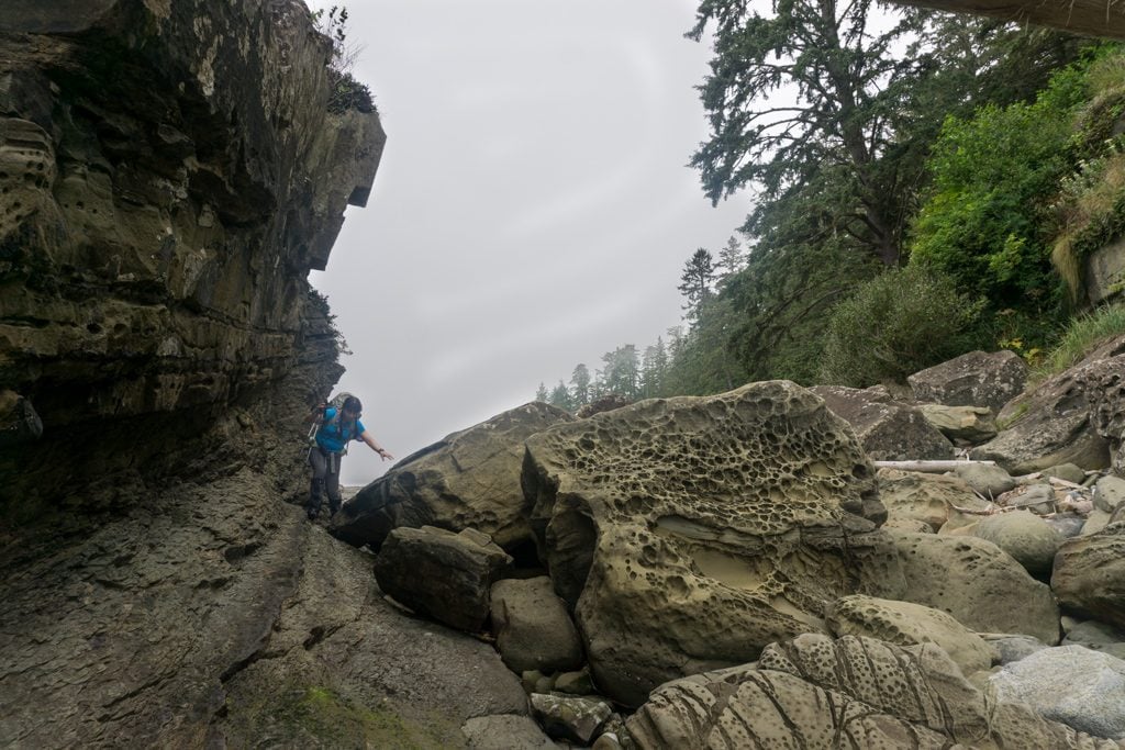 A hiker scrambles over rocks at a headland on the West Coast Trail