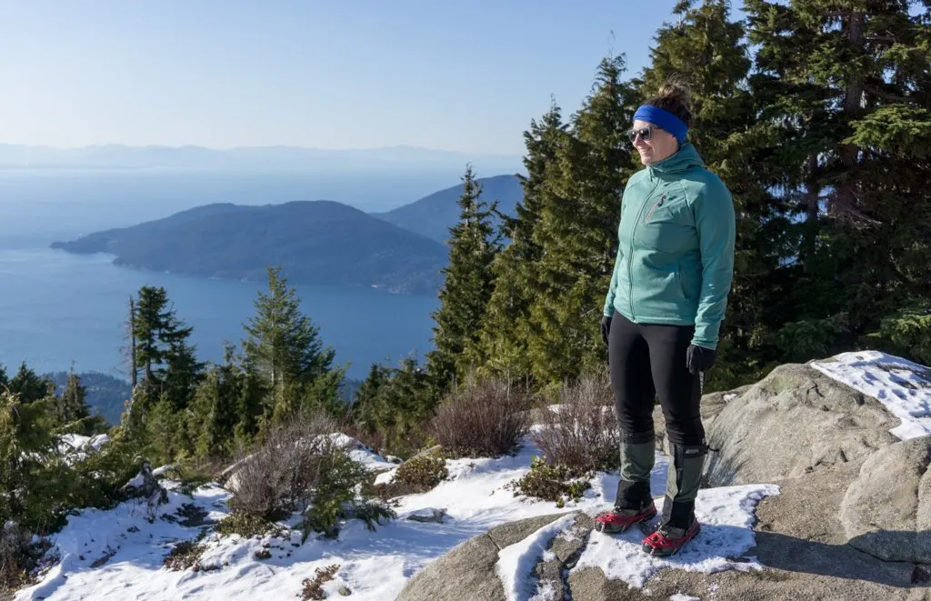 A hiker stands on top of a rocky bluff in the snow wearing microspikes