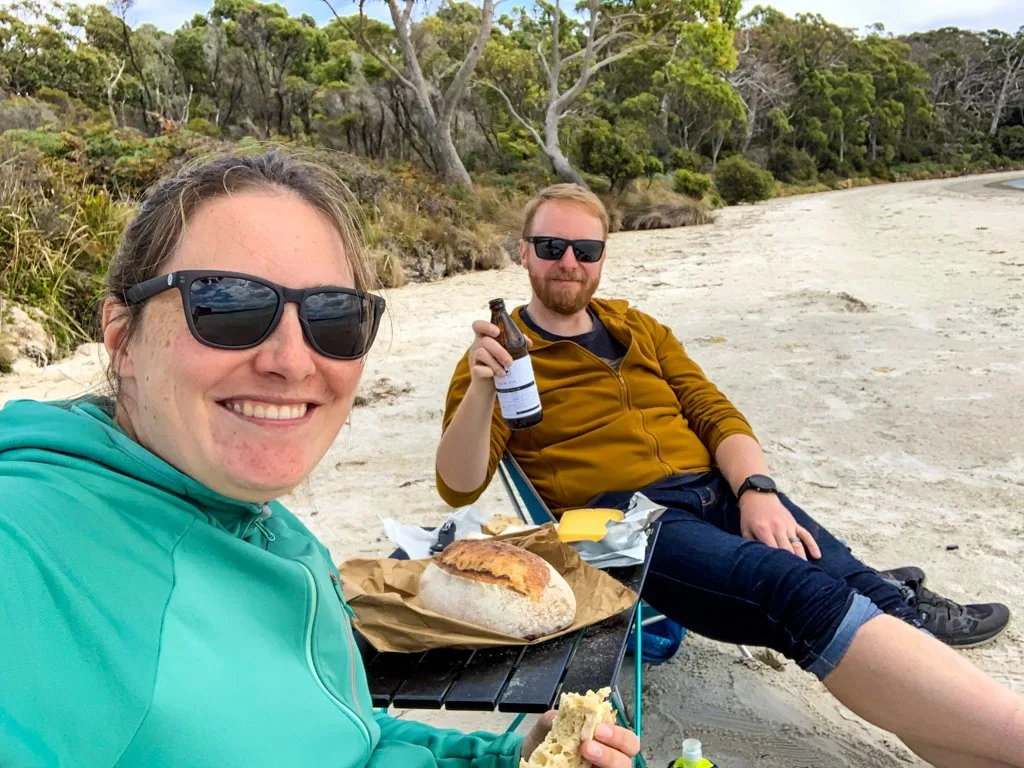 A man and a woman eat bread and cheese on a sandy beach