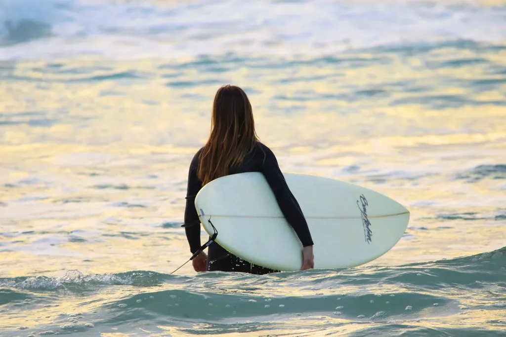 A woman in a wetsuit walks into the water holding a surfboard
