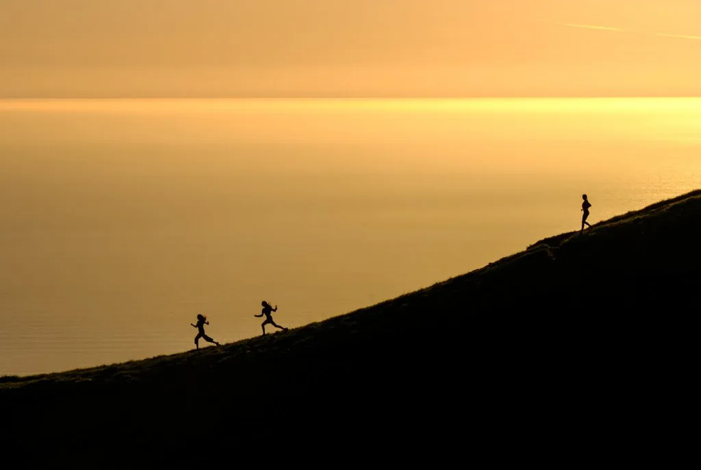 The silhouettes of three women as they run down a hill at sunset. Get this list of women's adventure films about running.