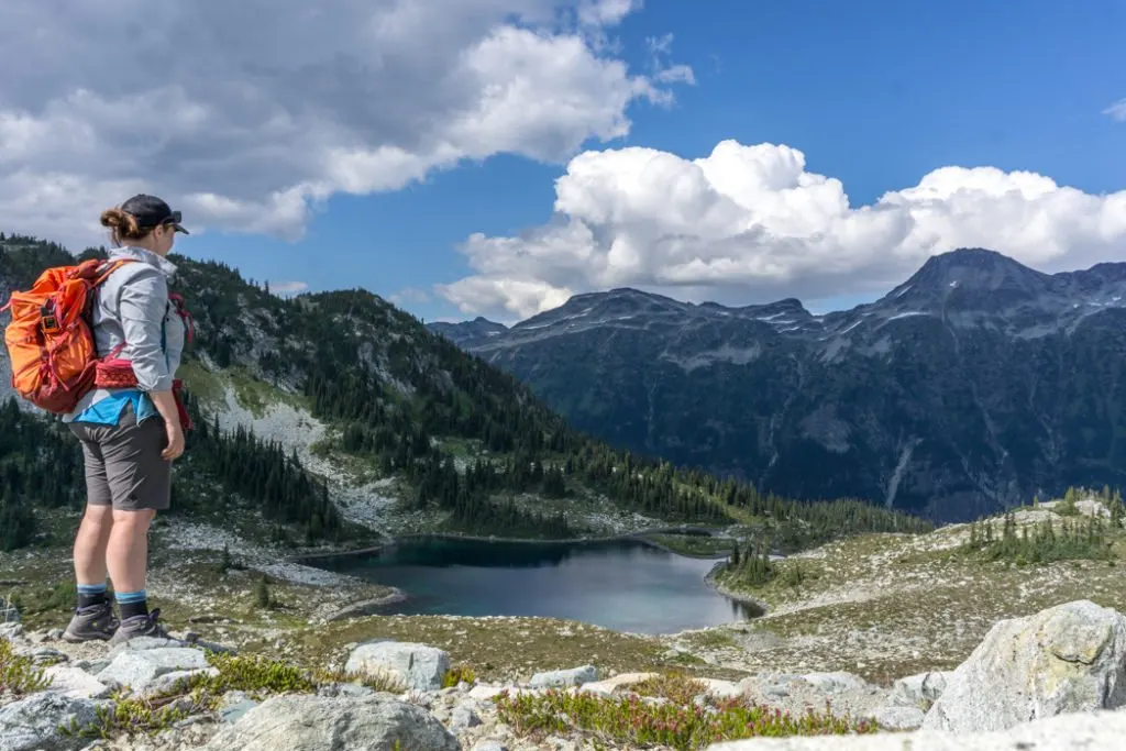 A woman on a backpacking trip standing in front of an alpine lake with mountains in the background