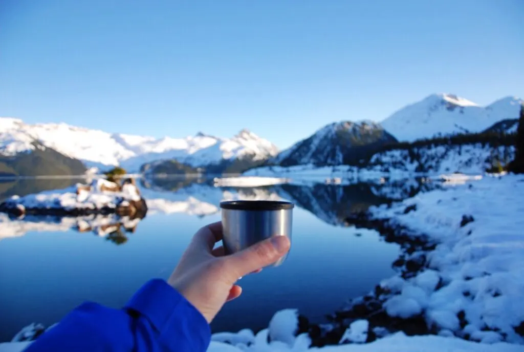 A snowshoer holds a cup of hot chocolate from a thermos in front of a snowy lake