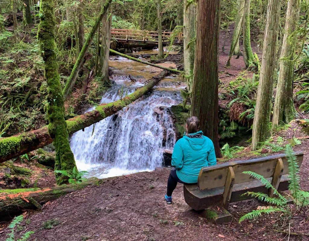 A woman sits on a bench and looks at a waterfall in Cliff Gilker Park on the Sunshine Coast, BC