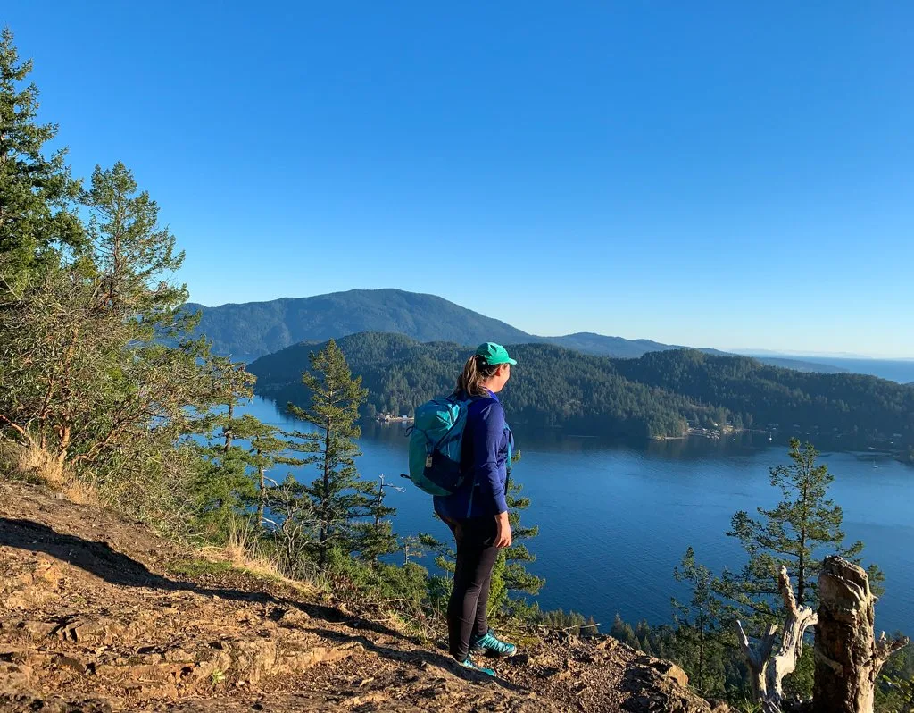 A woman with a bacpack stands at the edge of a cliff with a view of the ocean and islands on Soames Hill Trail on the Sunshine Coast, BC