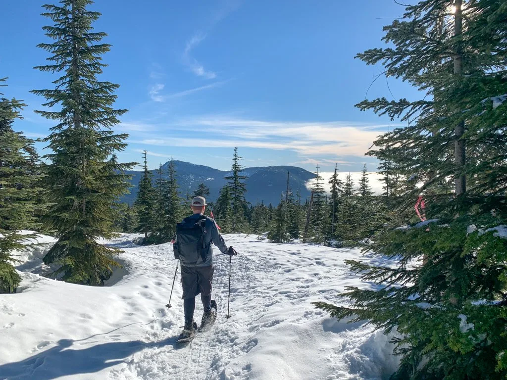 A man snowshoeing towards an opening in the trees with a view of the ocean at Dakota Ridge on the Sunshine Coast, BC