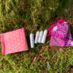 Menstrual products including pads, tampons and a cup rest on a mossy forest floor. Learn how to camp and hike on your period
