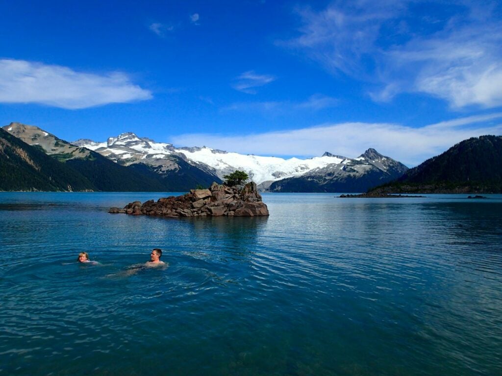 Two hikers swimming in Garibaldi Lake with glaciers in the background