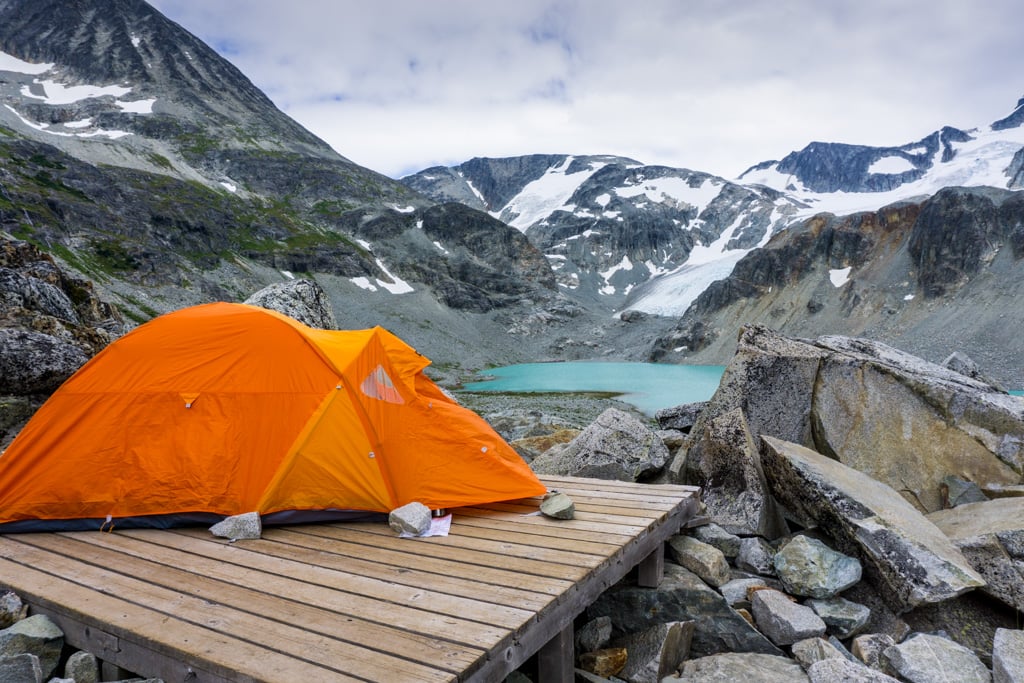 An orange tent sits on a wooden tent platform in front of a blue lake and a mountain with glaciers. Backcountry camping at Wedgemount Lake in Garibaldi Provincial Park.