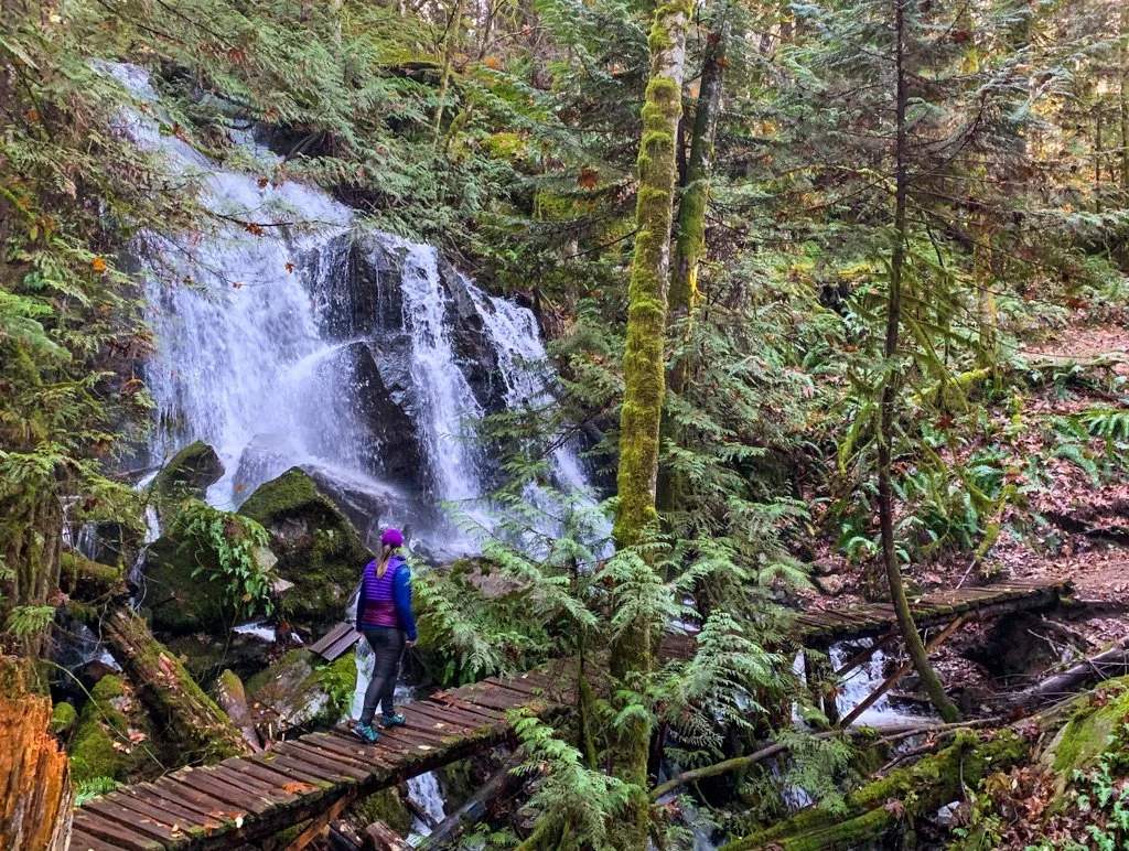 A female hiker wearing hiking tights crosses a bridge in front of a waterfall.