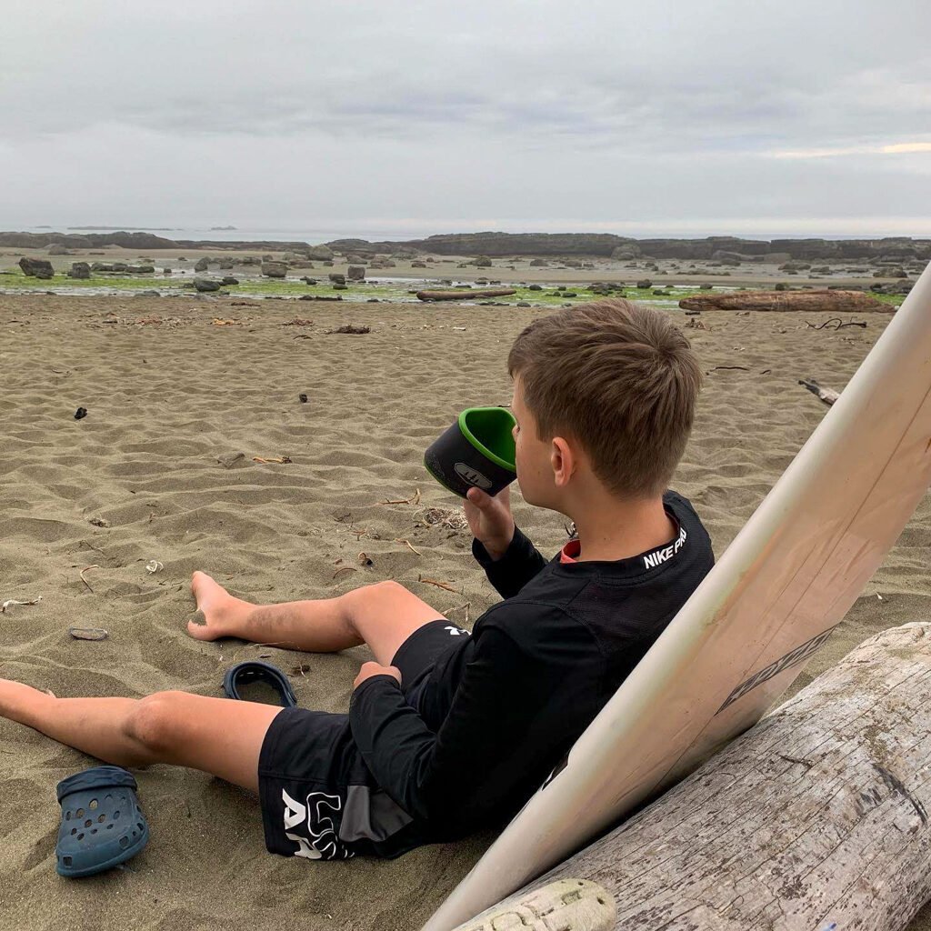 A boy drinking from a mug on a beach on the West Coast Trail