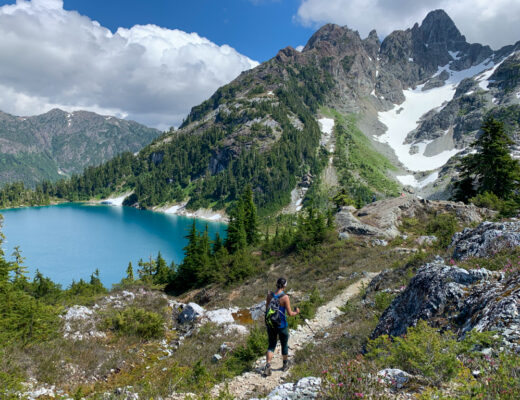 A hiker walks on a trail with a lake and snowy mountain in the background. Use this hiking glossary to get definition sofr hiking terms