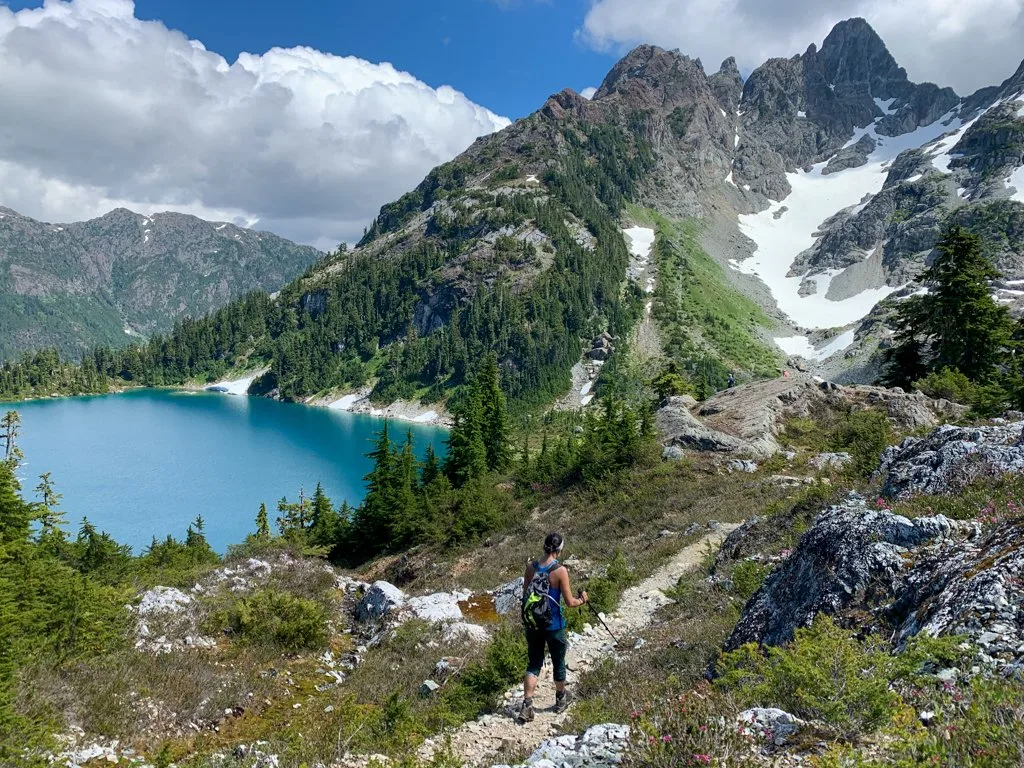 A hiker walks on a trail with a lake and snowy mountain in the background. Use this hiking glossary to get definition sofr hiking terms