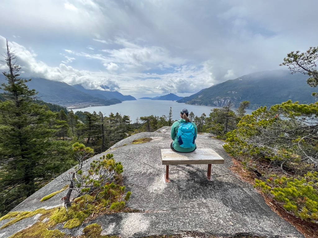 A female hiker wearing a blue backpack sits on a bench on a granite outcropping looking at a view of the ocean at Murrin Park near Squamish.
