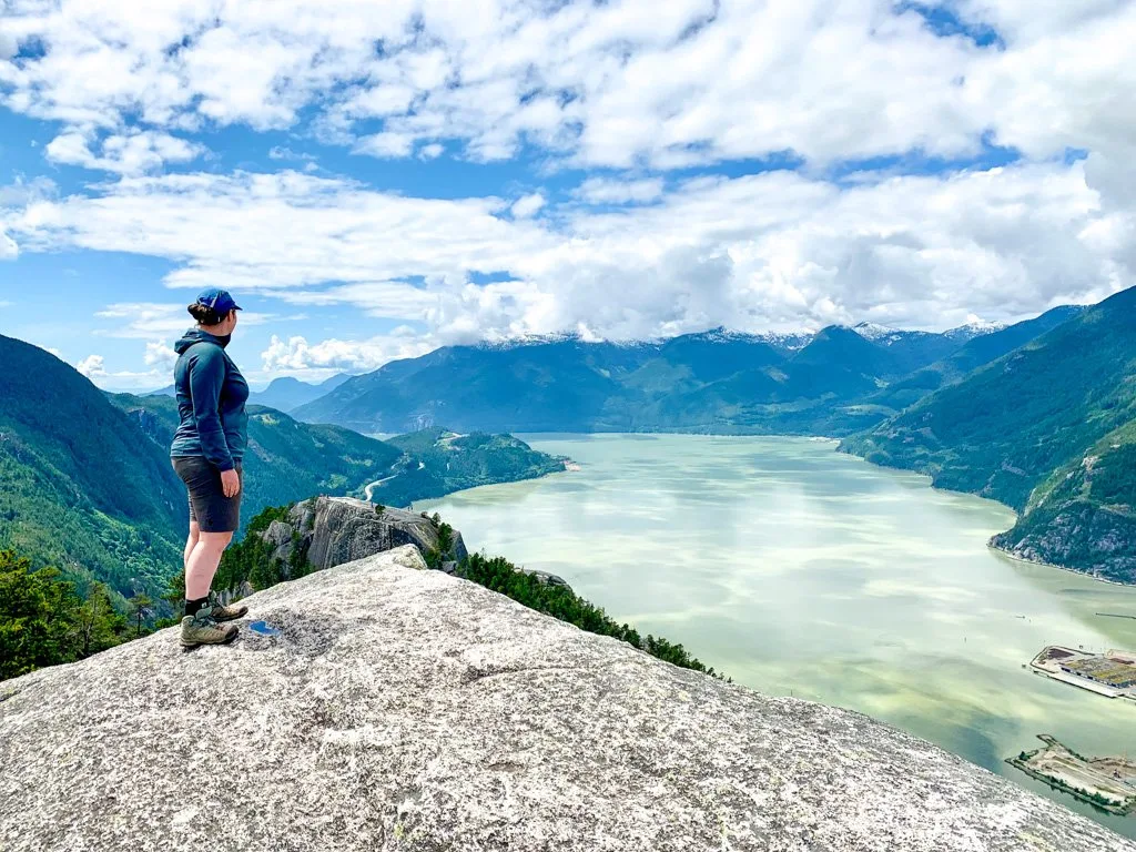 A female hiker wearing a blue fleece and shorts stands on top of the Stawamus Chief, a hiking trail in Squamish, BC