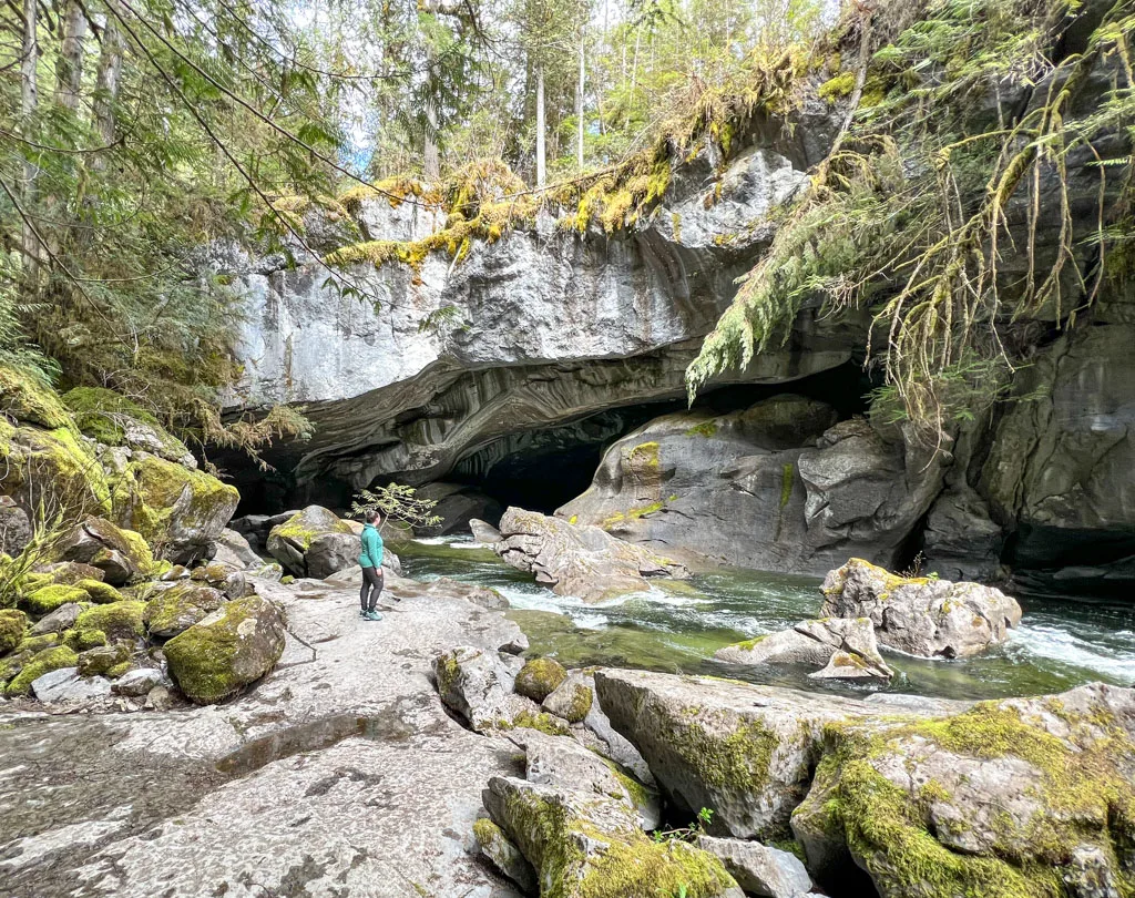 A woman in a green top stands at the entrance to Little Huson Cave on Northern Vancouver Island. The creek is flowing into a huge cave surrounded by evergreen trees and moss.