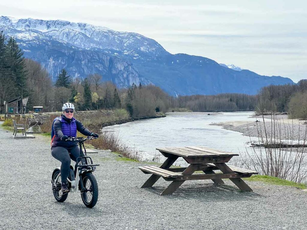 A woman rides the Rad Power RadMini Step-Thru electric bike on the dyke in Squamish with snow on the mountains in the background