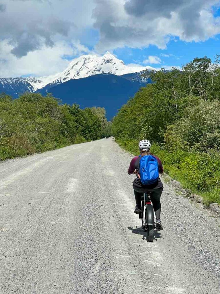 A woman wearing a backpack rides the Rad Power RadMini Step-Thru on a gravel road with a snowy mountain in the background