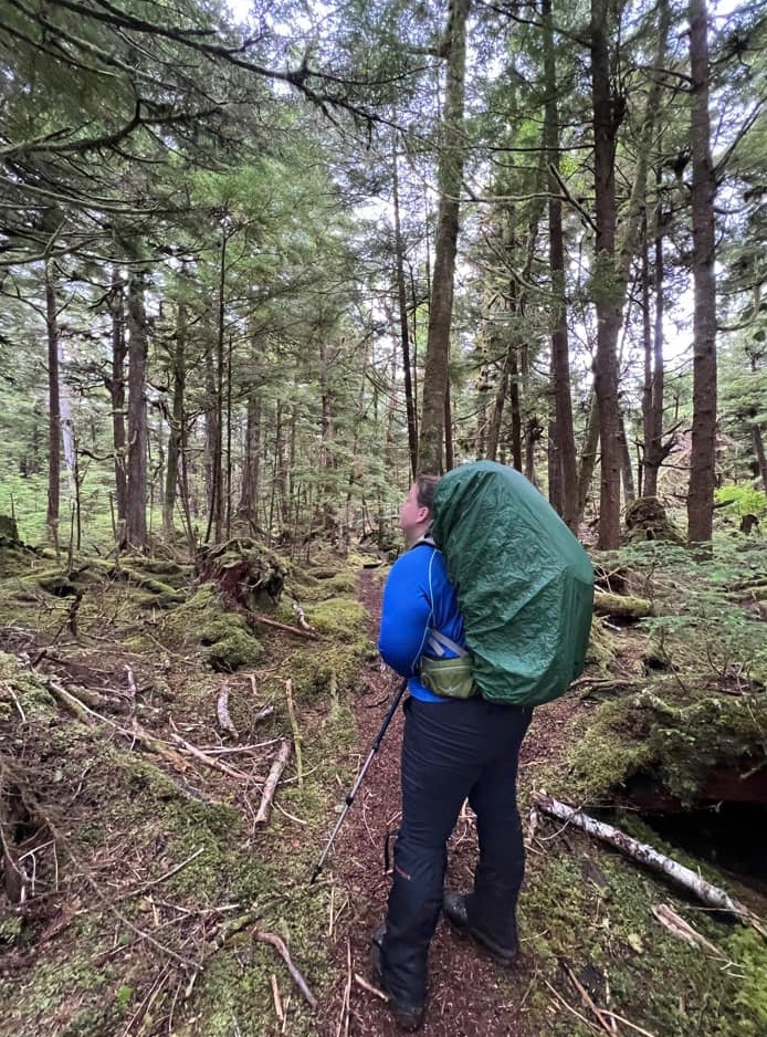 A woman wearing a big backpack with a rain green rain cover and Columbia Saturday Trail Stretch pants walks on a trail in a mossy forest