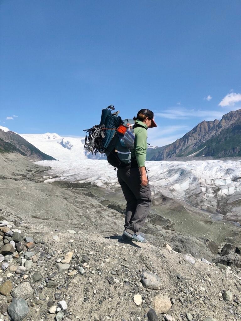 A woman wearing a backpack and REI Co-op Sahara Convertible Pants walks next to a glacier in Alaska