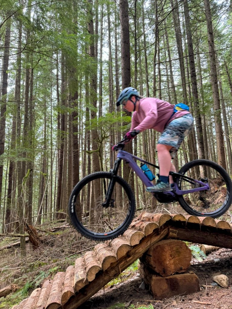 A woman mountain bikes over a ramp feature