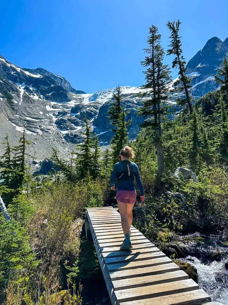 A hiker crosses a wooden bridge with mountains and glaciers in the background