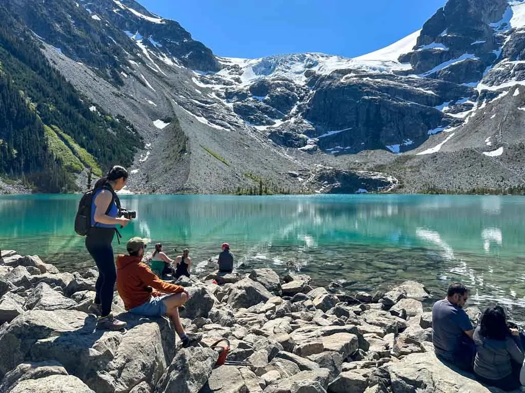 Hikers sit on rocks in front of a blue glacial lake with mountains in the background