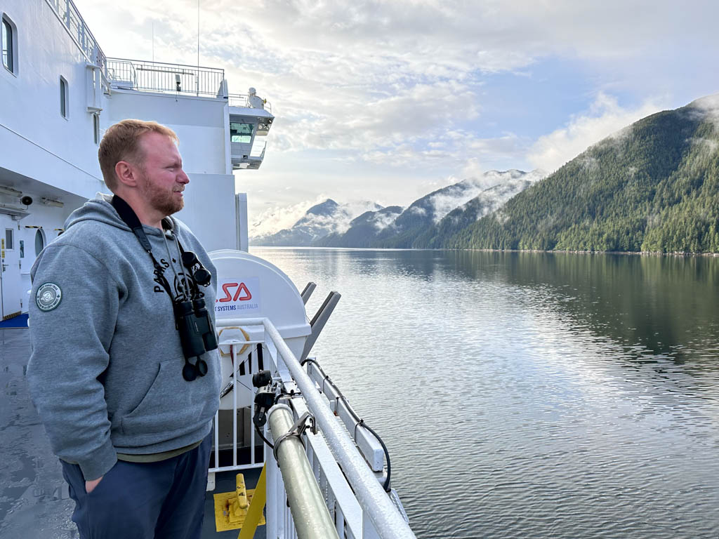 A man with binoculars around his neck looks at the scenery from the deck of the Inside Passage ferry