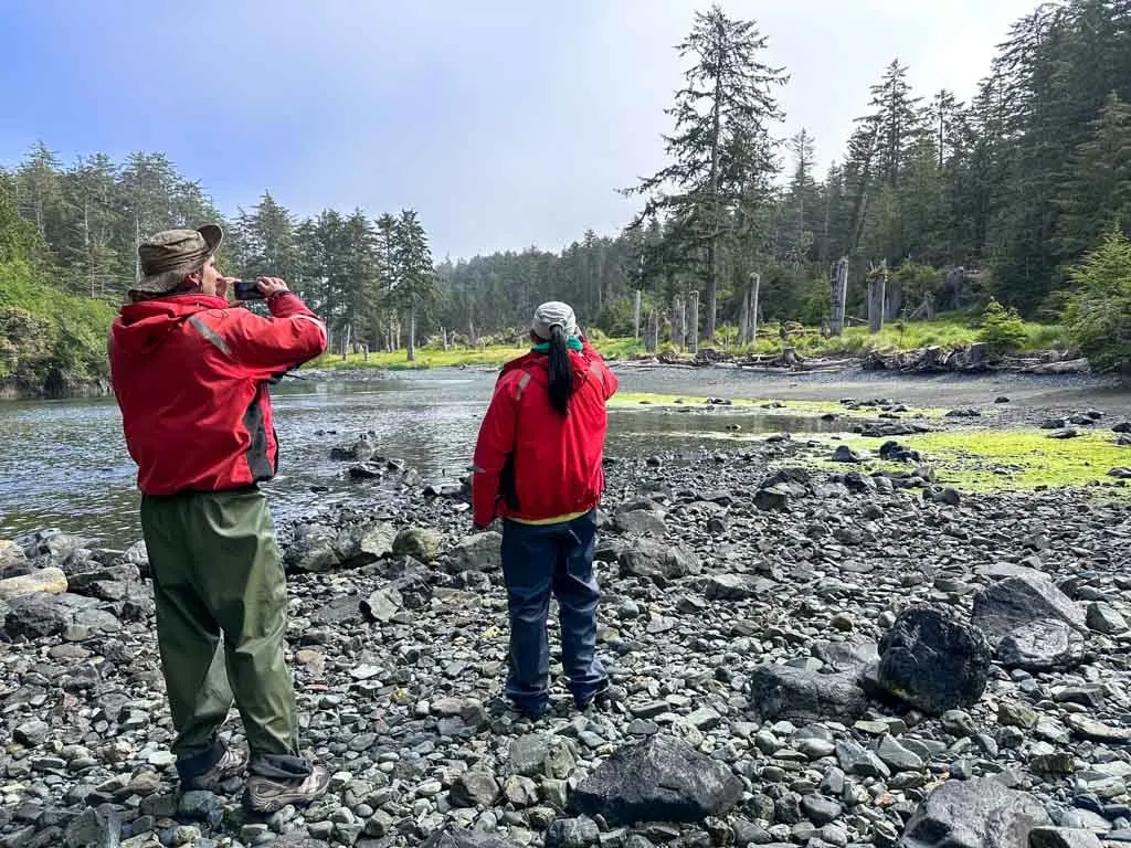 Two people wearing float coats and rain pants take photos at SGang Gwaay