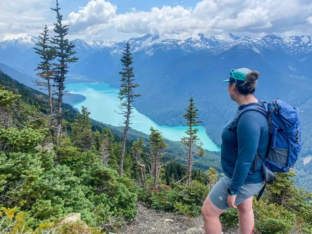 A female hiker wearing a backpack and a baseball cap looks down at Cheakamus Lake from the High Note Trail