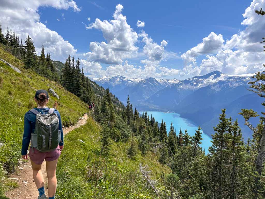 A hiker with a black backpack hikes along the High Note Trail with Cheakamus Lake in the background. 