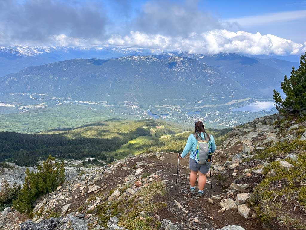 A female hiker wearing a backpack walks down the rocky High Note Trail with a view of Whistler valley below her