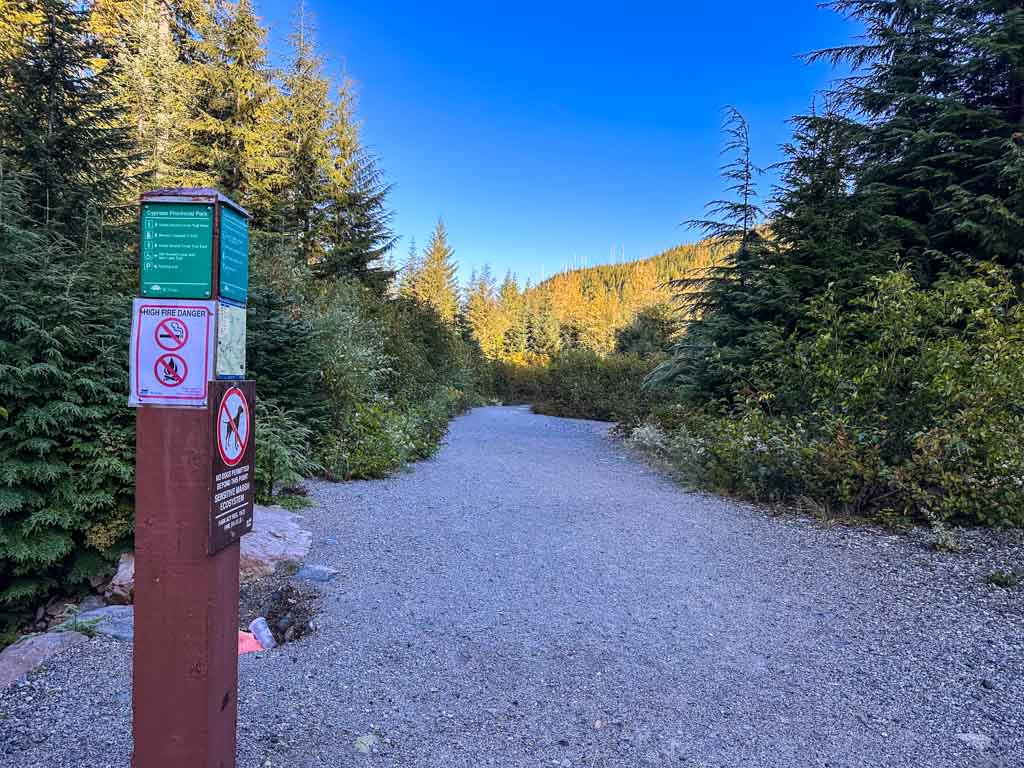 A sign post marks the junction between the Howe Sound Crest Trail to St. Mark's Summit and the Yew Lake Trail at Cypress Mountain Provincial Park. The sign post is on the side of a gravel road with tall coniferous trees on either side. 