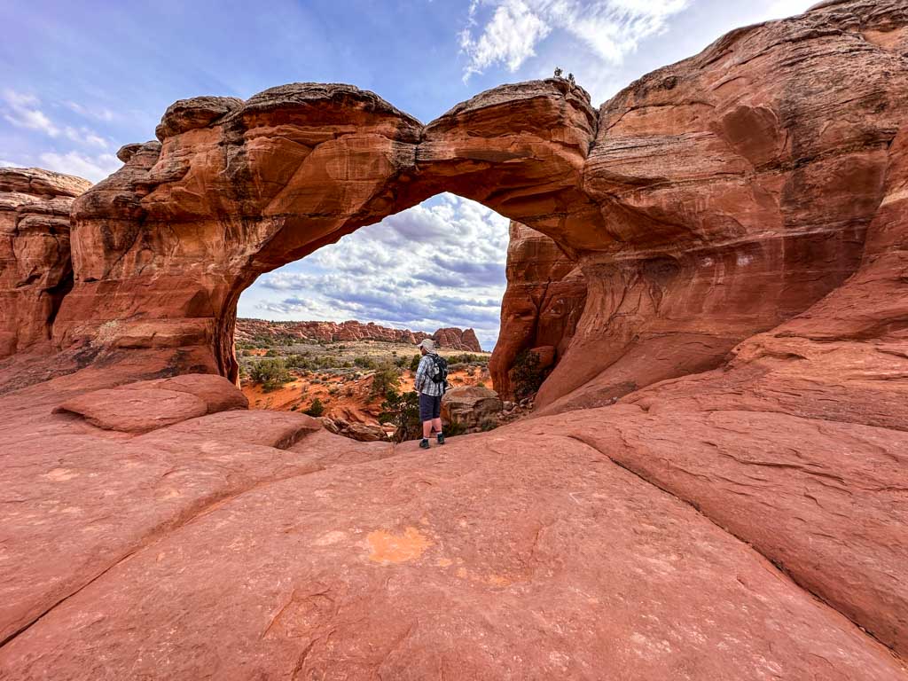 A hiker walks through Broken Arch in Arches National Park. You can see a crack in the top of the arch. 