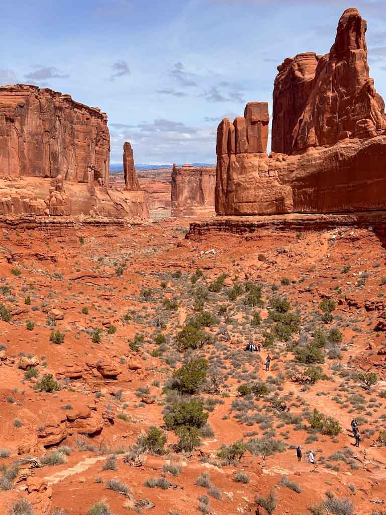 Looking north along the Park Avenue Trail in Arches National Park. There is a trail with hiker in the foreground. The trail passes between tall red rock spires. 