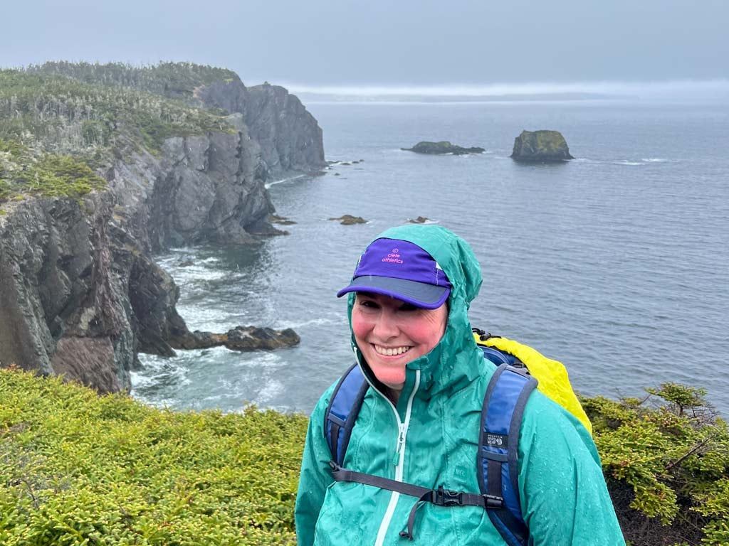 A woman wearing a rain coat and a backpack with a rain cover smiles on a rainy day on the Skerwink trail near Port Rexton