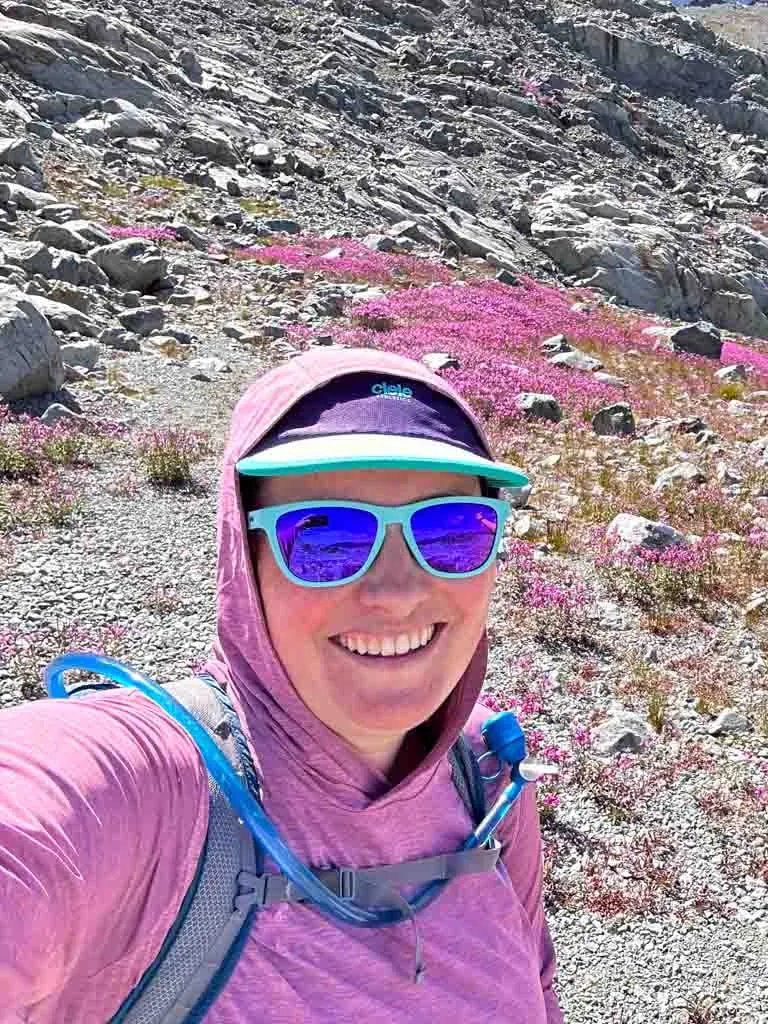 A hiker takes a selfie in front of alpine flowers wearing the Patagonia Capilene Cool Daily hoody with the hood up