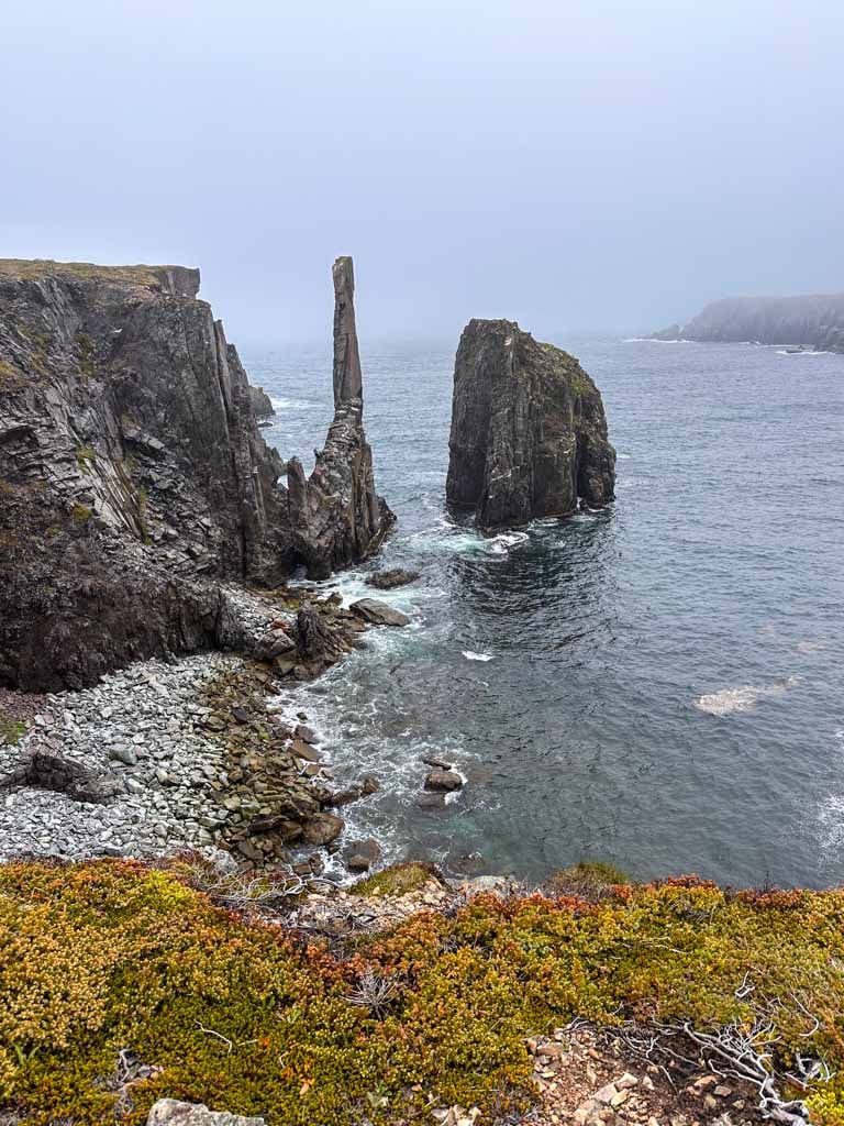 A tall and thin seastack called The Chimney rises out of the ocean in front of a foggy sky near Bonavista Newfoundland