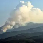 A plume of smoke rises above a forested mountain. Get tips for safe hiking in wildfire season