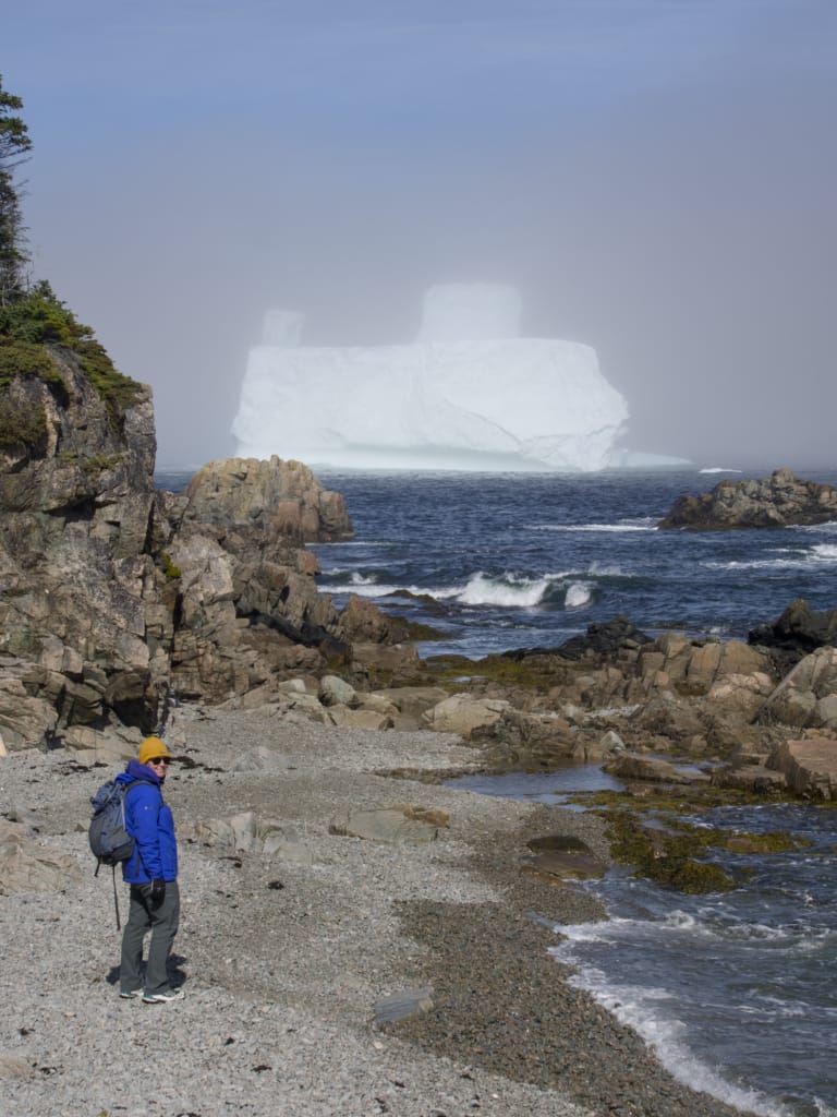 A hiker stands on a beach with a huge iceberg in the background near Twillingate Newfoundland