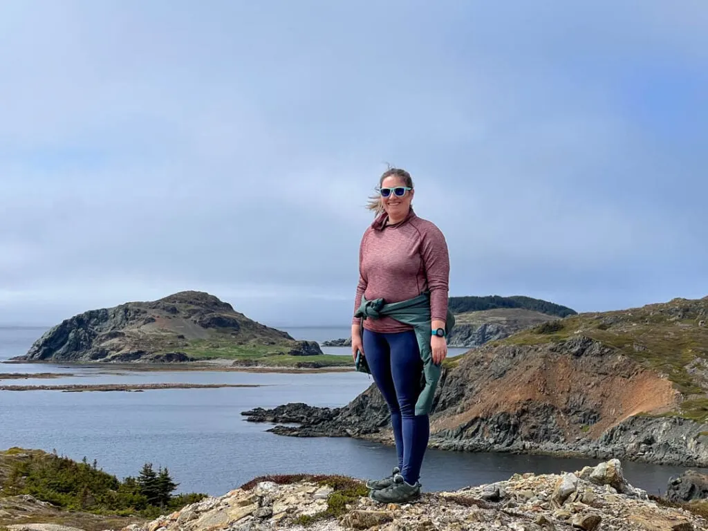 A woman poses on a hike in Twillingate. She is wearing a warm jacket tied around her waist. 