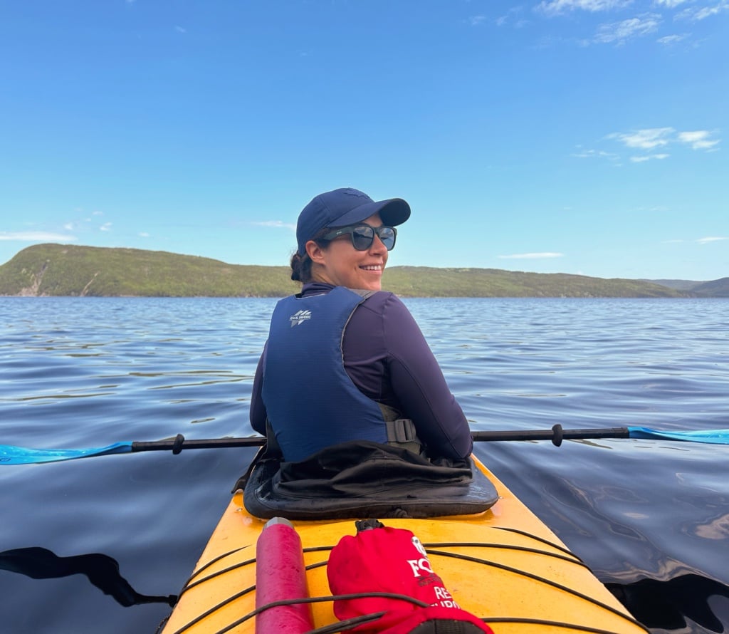 A woman turns to look at the camera from the front seat of a double kayak. In the background you can see the ocean and an island in the Bay of Islands near Corner Brook, Newfoundland