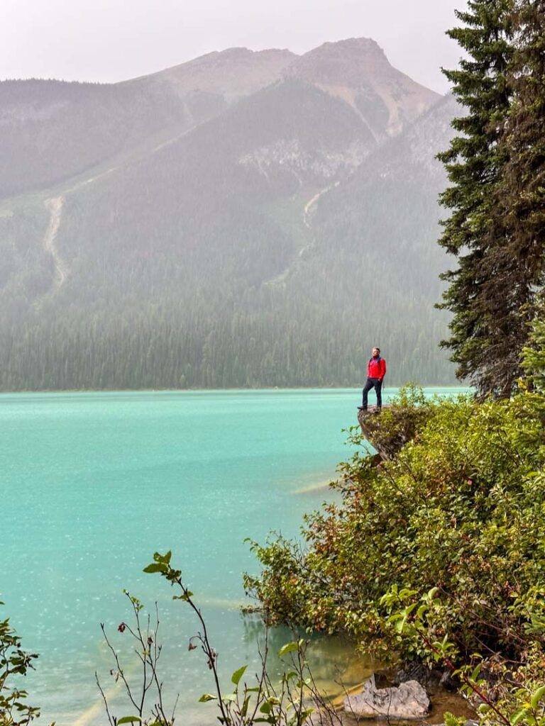 A man in a red jacket stands on the shores of Emerald Lake in the pouring rain. 