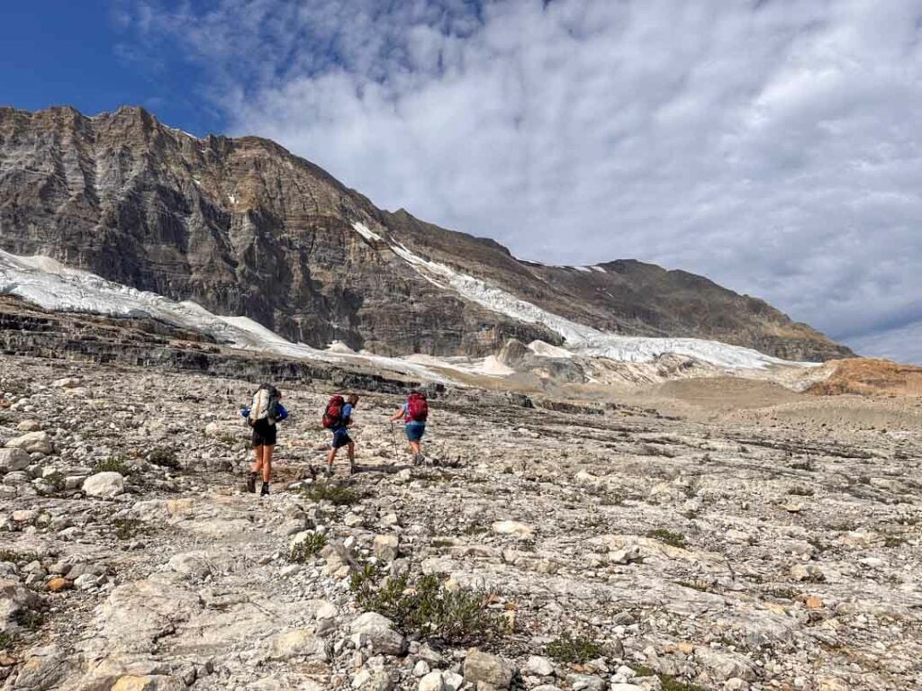 Three hikers with large backpacking packs on the Iceline Trail (part of the Yoho Valley Loop) in Yoho National Park. They are walking through a rocky landscape with glaciers on the mountain behind them.