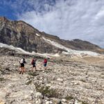 Three hikers with large backpacking packs on the Iceline Trail (part of the Yoho Valley Loop) in Yoho National Park. They are walking through a rocky landscape with glaciers on the mountain behind them.